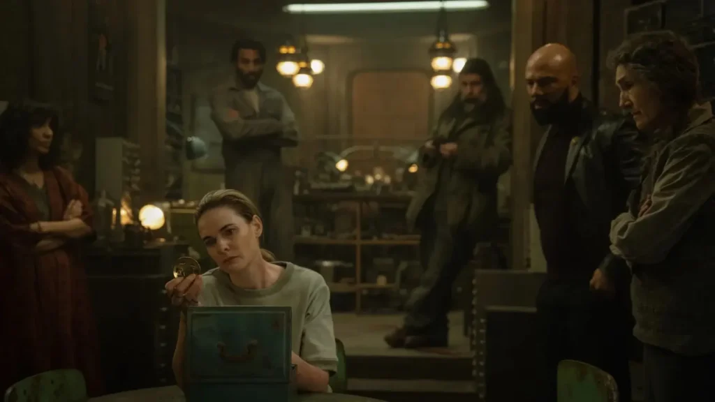 A scene from Silo featuring Juliette seated at a table examining a round relic from a metal box, surrounded by a group of serious-looking characters in a dimly lit workshop.