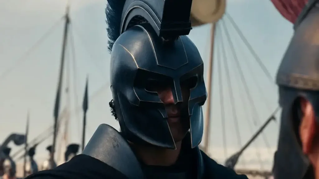 5 Great Greek Mythology Movies to Watch After ‘The Odyssey’ A close-up view of a warrior in the open, clad in dark armor and an equally ornate helmet topped with a blue crest. Behind him one can see the masts and ships of what is clearly an outdoor setting.