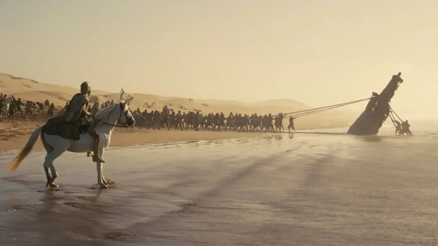 Has Christopher Nolan Really Revolutionized Fantasy? The Technologies That Made The Odyssey Unique A scene from The Odyssey showing an armored rider on a white horse watching a long line of men pull a giant wooden horse ashore with heavy ropes on a sandy beach.