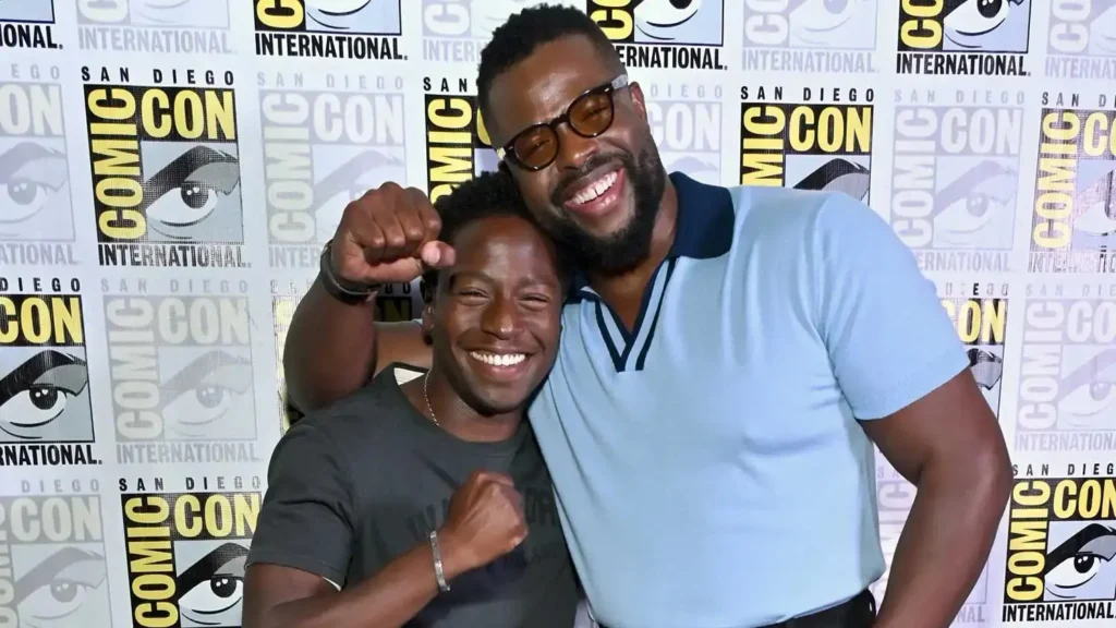 Actors David Jonsson and Winston Duke smiling cheerfully together in an embrace with raised fists in front of a San Diego Comic-Con press backdrop.