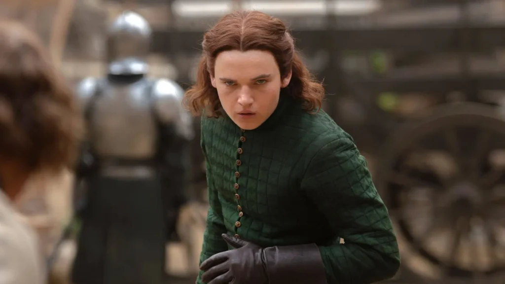 A young man with shoulder-length reddish-brown hair wearing a quilted green tunic and leather gloves leaning forward with a fierce stare in House of the Dragon.