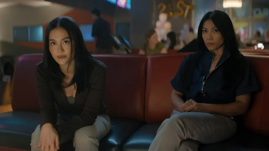 Lila Hoth and another woman sitting on a leather bench inside a bowling alley or entertainment venue looking towards the camera in Reacher.