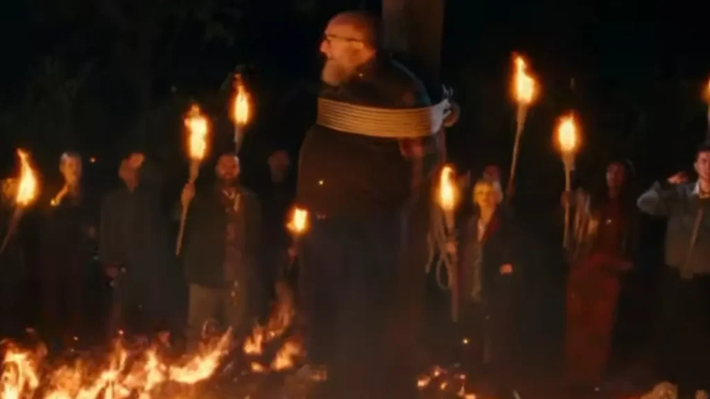 Brian Posehn tied to a wooden stake surrounded by glowing flames and a crowd holding burning torches at night.