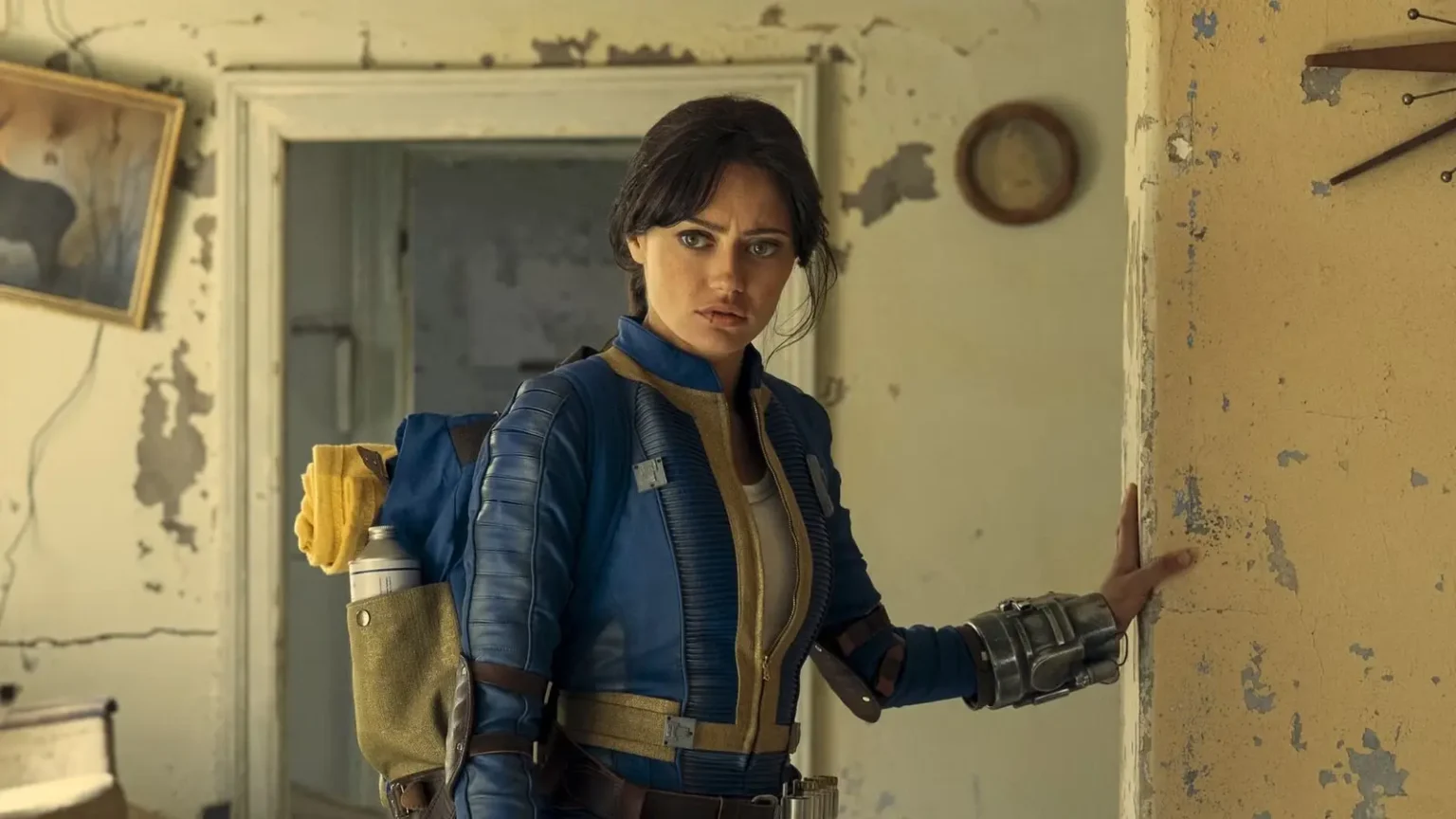 Do New Fallout Season 3 Set Photos Hint at the Brotherhood’s Deepest Lore? Ella Purnell as Lucy MacLean wearing a blue Vault jumpsuit and Pip-Boy, standing cautiously in a dilapidated house in Fallout.