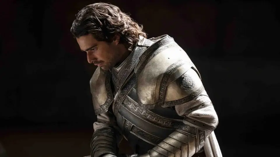 Ser Criston Cole in full Kingsguard armor looking downward with a somber expression against a dark background in House of the Dragon.