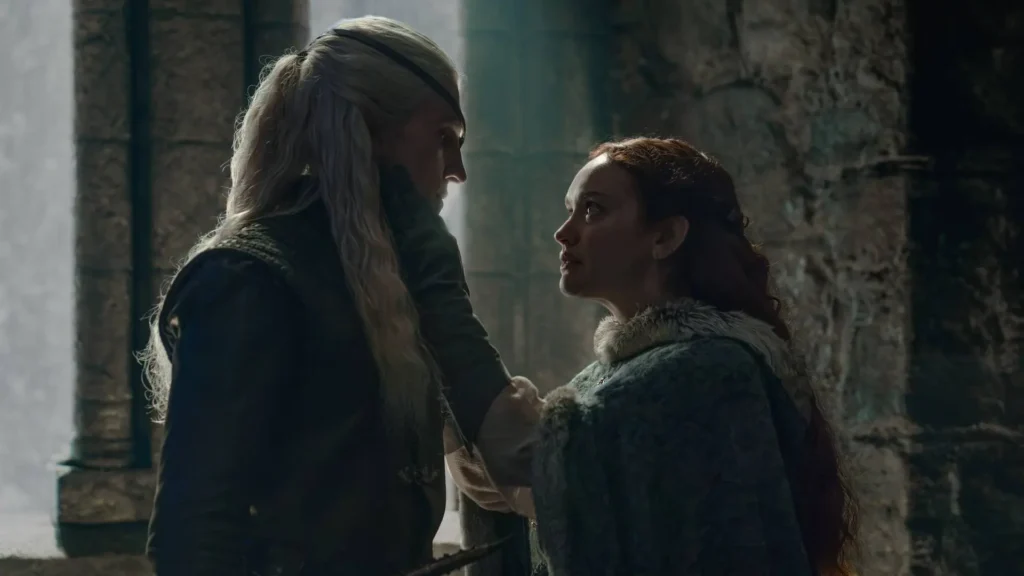 Ewan Mitchell as Aemond Targaryen touching the cheek of Olivia Cooke as Alicent Hightower in a shadowy stone room in House of the Dragon.