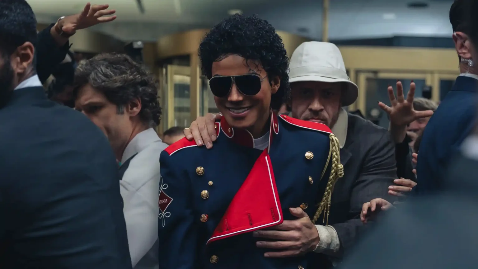 Jaafar Jackson as Michael Jackson smiling while wearing dark aviator sunglasses and a military-style jacket surrounded by fans and security.