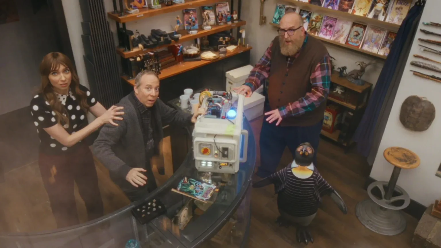 Kevin Sussman, Lauren Lapkus, Brian Posehn, and a penguin puppet reacting in shock to a glowing sci-fi device in a comic book shop.