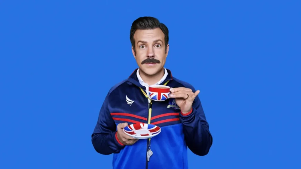 Ted Lasso: The Complete Story So Far Ahead of Season 4 Jason Sudeikis as Ted Lasso holding a Union Jack teacup and saucer against a bright blue background in Ted Lasso.