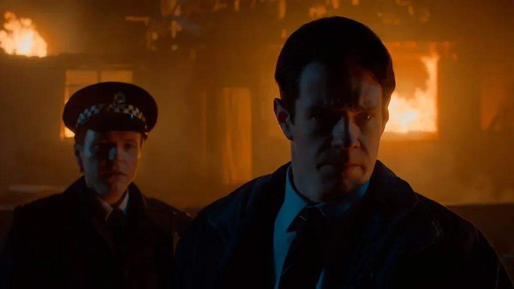 Connor Swindells as Detective Ed McCusker looking somber in front of a burning building with a uniformed officer behind him in Netflix's The Bombing of Pan Am 103.