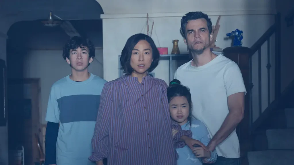 The Last House Review: A Great Premise That Loses Its Way Greta Lee, Wagner Moura, and two children looking forward with shocked, fearful expressions in a dimly lit home.