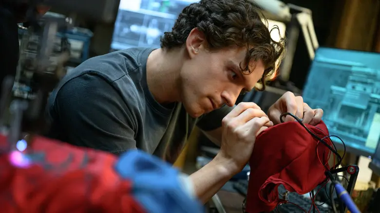 Tom Holland as Peter Parker leaning over a workbench and carefully working on his red Spider-Man suit.